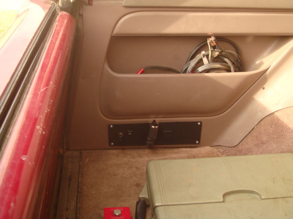 Interior view of a vehicle showing a storage pocket with jumper cables and a 12v power outlet.