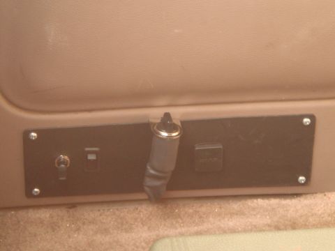 1992 Ford Bronco - A close-up view of a black panel mounted on a tan surface, featuring a toggle switch, a rectangular outlet labeled "12V OUTLET", and a silv…