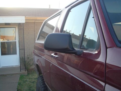 1992 Ford Bronco - The side profile of a maroon Ford Bronco is visible, parked next to a house with a white door and brick siding. The vehicle's side mirror a…