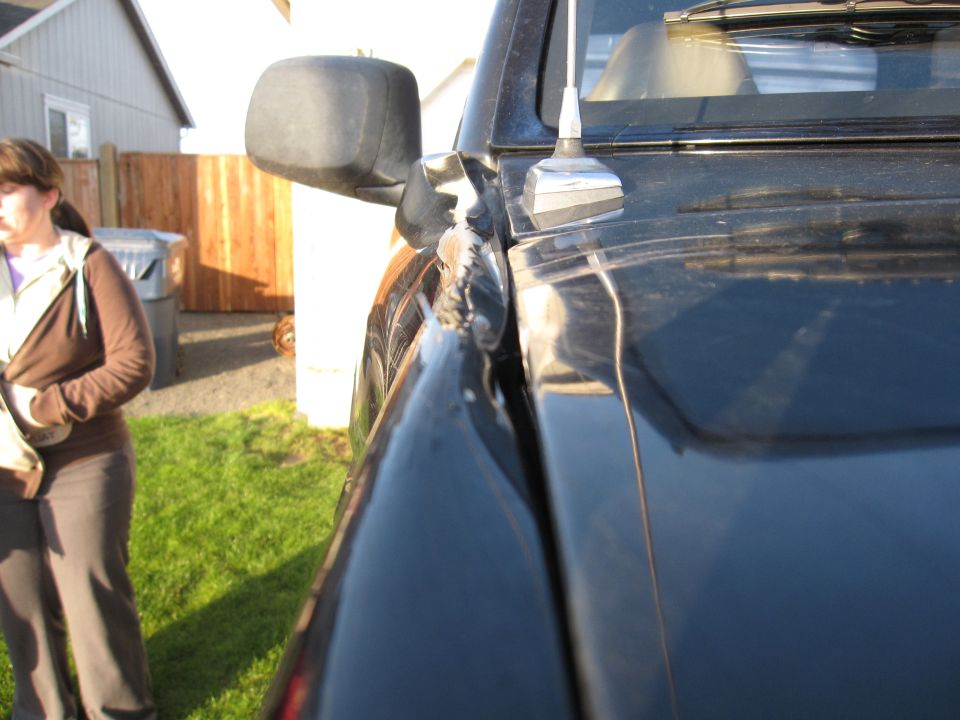 Black vehicle side mirror and hood detail, woman in background.