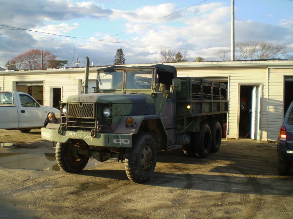 Camouflage military truck parked in front of a building with a white pickup truck to its left.