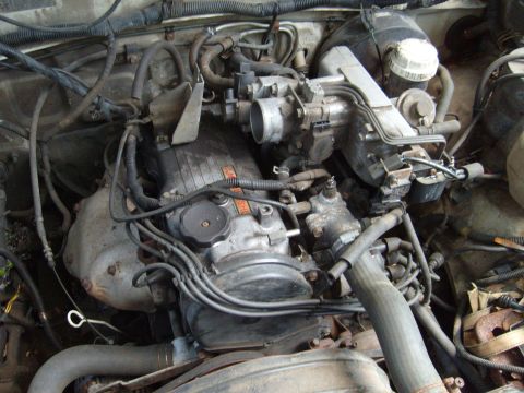 1992 Ford Bronco - The image shows a close-up view of a dirty, complex engine bay with many hoses, wires, and engine components visible. A silver-colored engi…