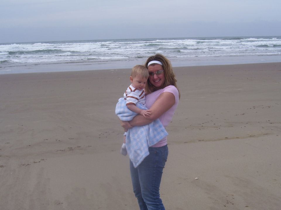 Woman holding a baby on a sandy beach with waves in the background. Watermark reads SUPERMOTORS