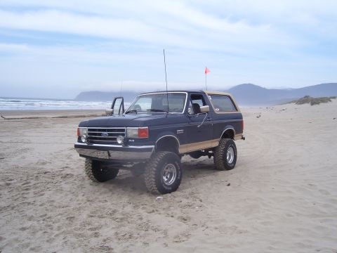 1991 Ford Bronco - This lifted Ford Bronco is parked on the sand dunes with the ocean in the background under a cloudy sky.