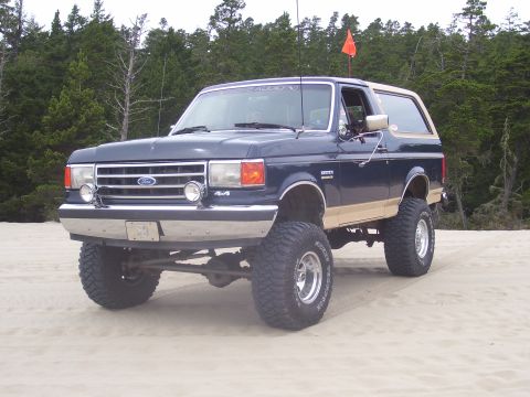 1991 Ford Bronco - A lifted 1991 Ford Bronco with tan trim sits on a sandy terrain, with trees in the background.