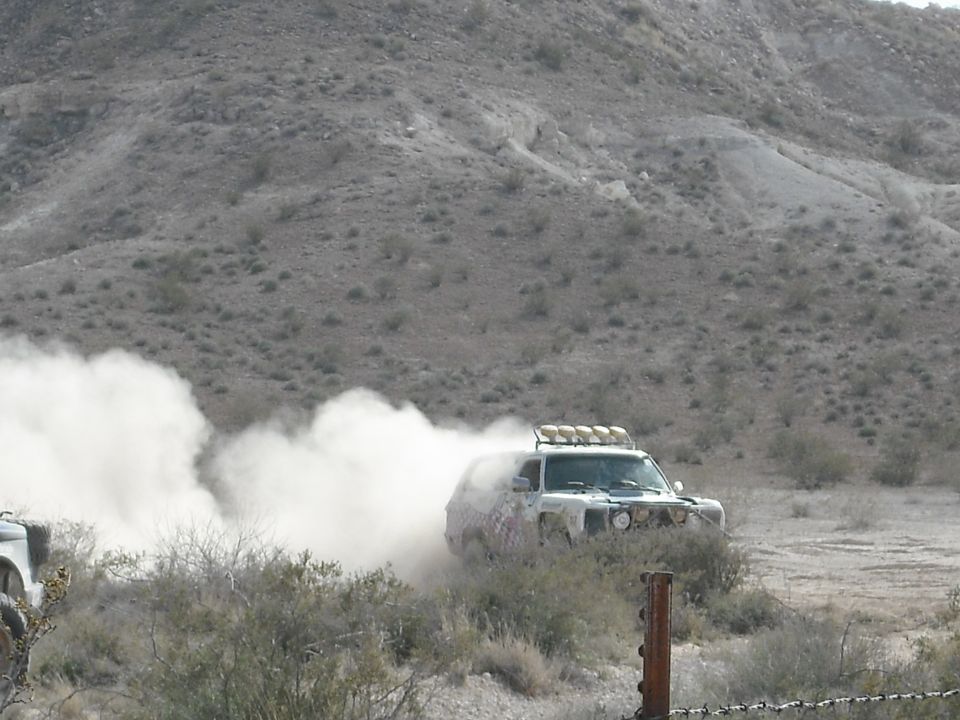 Off-road truck with light bar on roof kicking up dust on a dirt track. Another vehicle is partially visible to the left