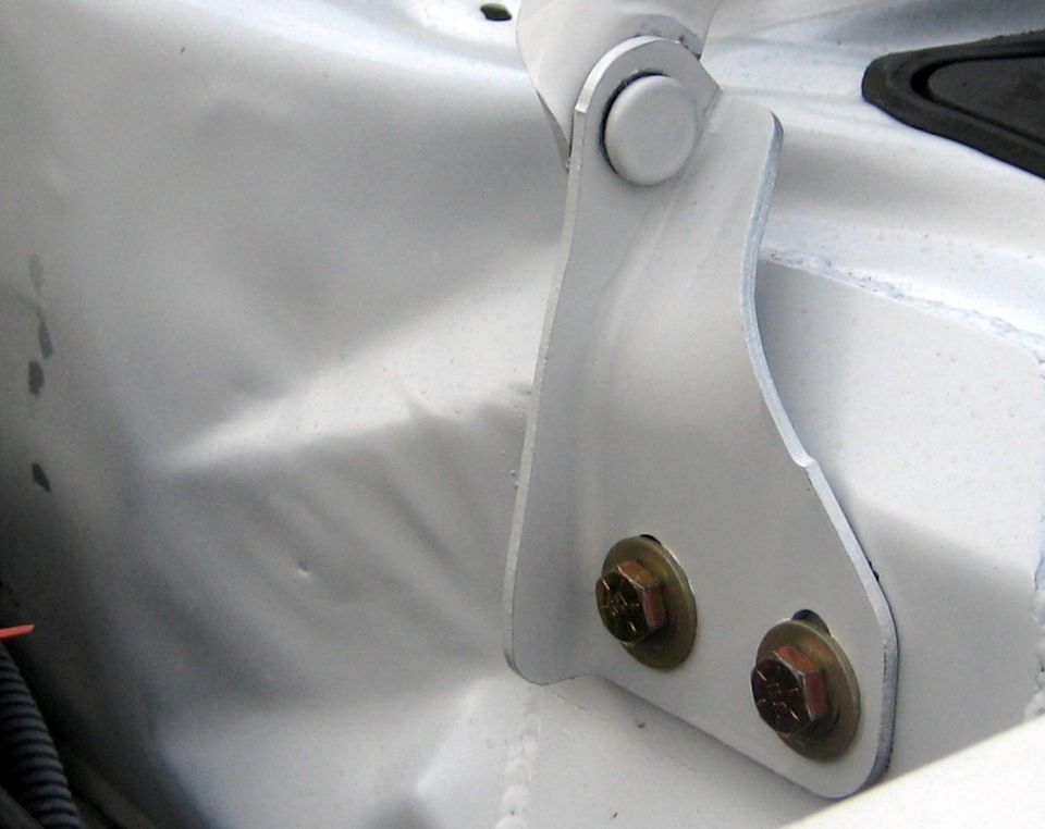 White painted metal hood hinge bracket with two hex bolts attached to a dented white metal panel