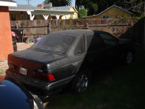 1989 Ford Taurus - The rear of a black sedan is visible, parked in a grassy area with houses in the background.