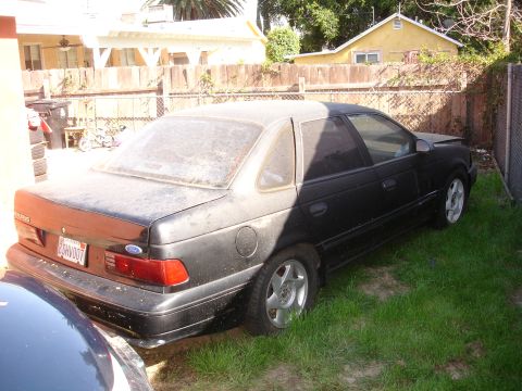 1989 Ford Taurus - A dusty black 1989 Ford Taurus sedan is parked in a grassy yard, viewed from the rear.