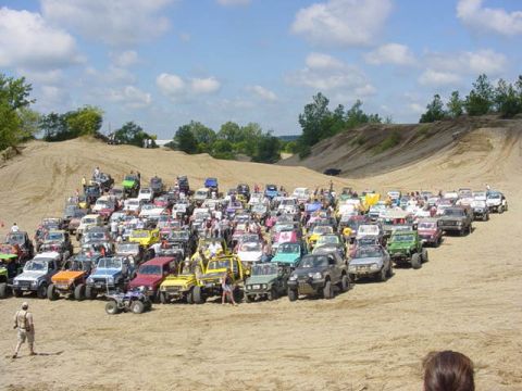 1992 Ford Bronco - A large gathering of off-road vehicles are parked on a sandy terrain under a partly cloudy sky.