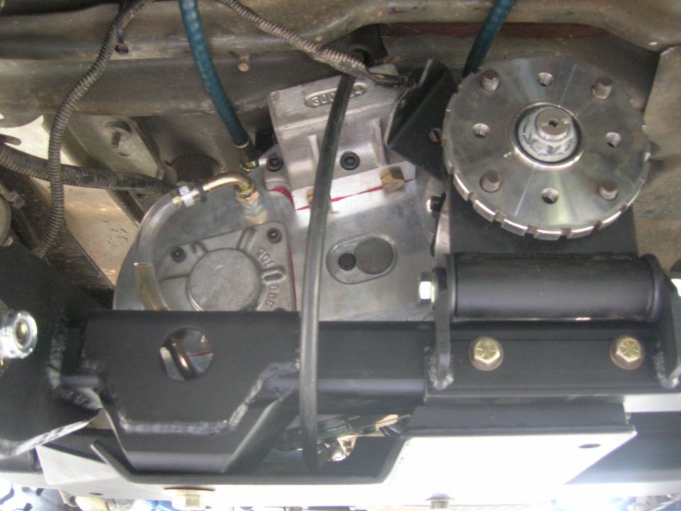 Underside view of vehicle transmission with metal casing, hoses, and a circular component with bolts