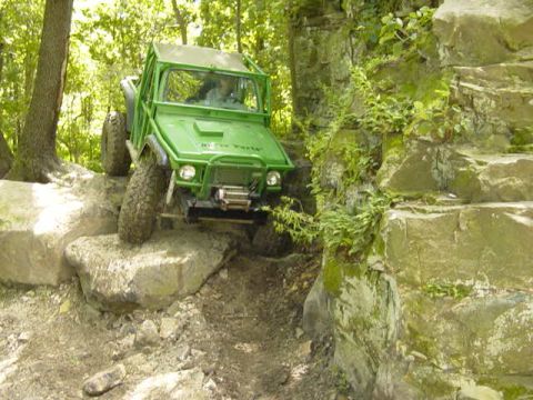 1992 Ford Bronco - A green off-road vehicle navigates a challenging rocky terrain, with trees and foliage in the background.