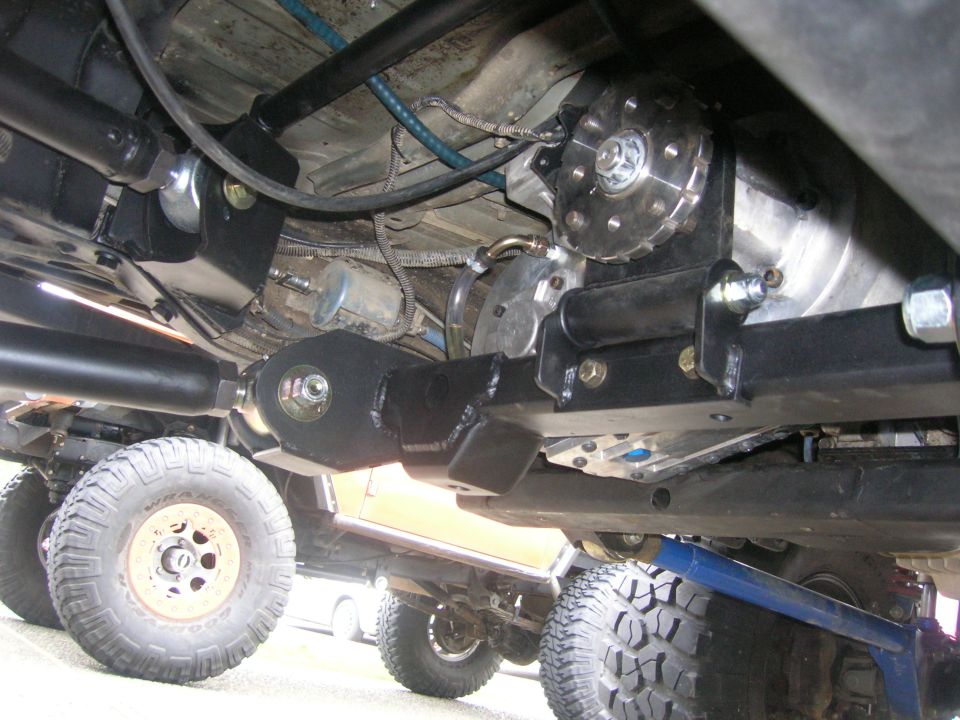 Undercarriage view of a modified off-road vehicle showing suspension components and tires. Another vehicle is partially visible