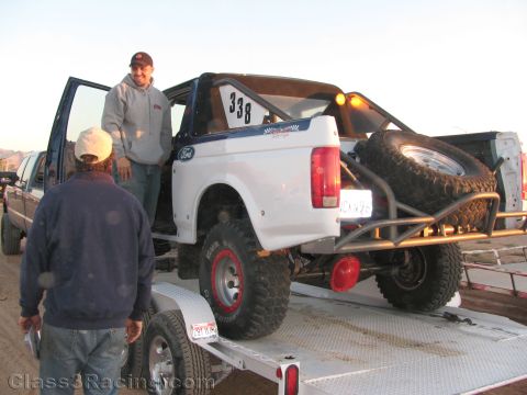 1990 Ford Bronco - white Ford off-road race truck numbered 338 with spare tire sits on a flatbed trailer as two men stand beside it at dusk.
