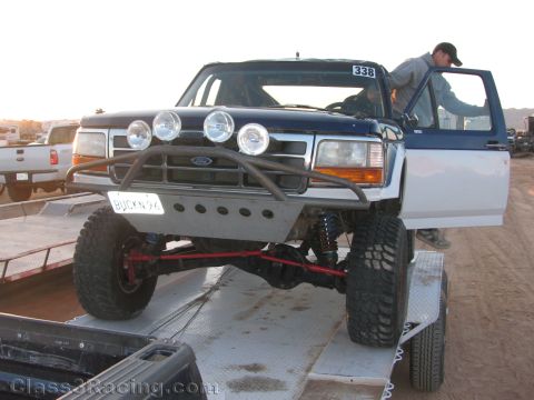 1990 Ford Bronco - blue and white Ford off-road race truck numbered 338 with roof light bar is parked on a flatbed trailer in a dirt lot.