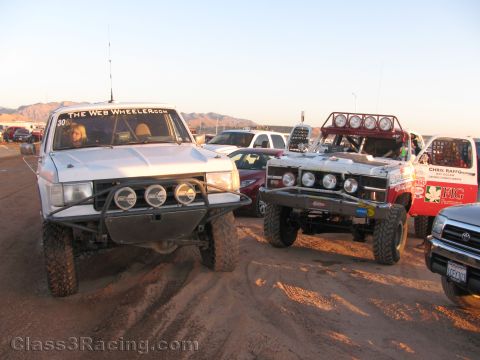 1990 Ford Bronco - silver off-road truck lettered The Web Wheeler and a red number 304 truck sit among other race vehicles in a dusty desert lot.