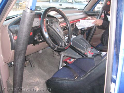 1990 Ford Bronco - Interior of an off-road race truck showing a padded roll cage, Ford steering wheel, single bucket seat and a person holding paperwork.