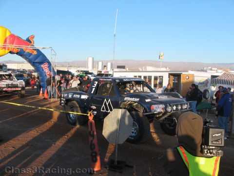 1990 Ford Bronco - dark-colored off-road race truck numbered 31 stages at a desert start line with spectators and traffic cones nearby.
