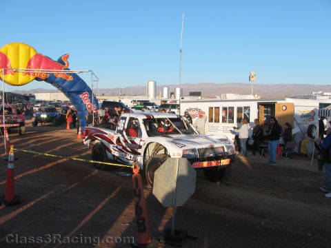 1990 Ford Bronco - white and red off-road race truck launches from a desert start line beside a Red Bull inflatable arch as a crowd watches.
