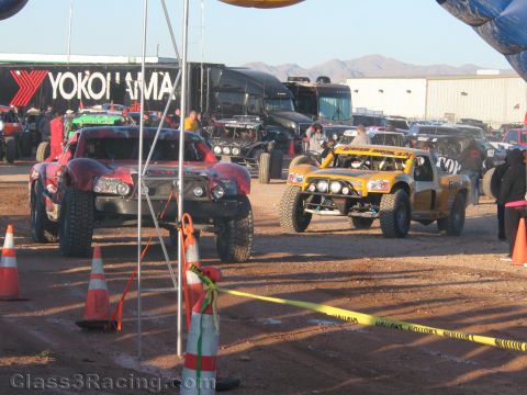 1990 Ford Bronco - red off-road truck and a yellow Fox-sponsored race truck sit among other vehicles in a dusty staging area with cones and tape.