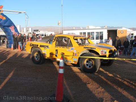 1990 Ford Bronco - yellow off-road race truck lettered Greer Bros Racing on Toyo Tires sits in a dusty desert staging area near a building.