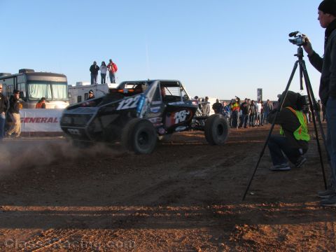 1990 Ford Bronco - black off-road race truck numbered 124 kicks up dust at a desert event while a cameraman films from a tripod and crowds watch.