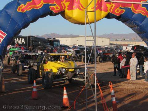 1990 Ford Bronco - yellow off-road buggy and other race vehicles stage under a Red Bull inflatable arch in a dusty desert lot with spectators.