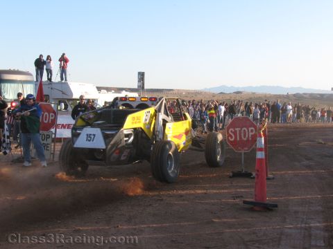 1990 Ford Bronco - yellow off-road buggy numbered 157 throws dirt as it launches past STOP signs at a desert race start with a crowd watching.
