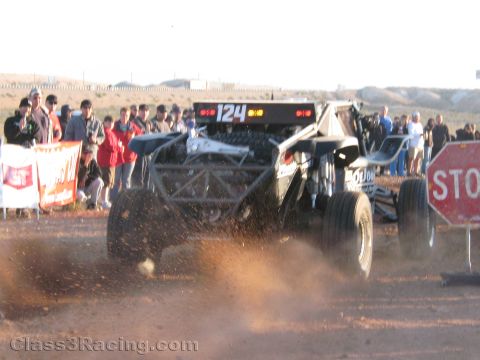 1990 Ford Bronco - off-road race buggy numbered 124 sprays a rooster tail of dirt as it accelerates past spectators and a STOP sign in the desert.