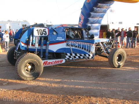 1990 Ford Bronco - blue off-road race buggy numbered 105 lettered Buchwitz Racing sits on BFGoodrich tires near a Red Bull arch in a dusty lot.