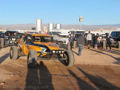 1990 Ford Bronco - orange and black off-road race buggy sits among other vehicles and people in a dusty desert staging area with mountains beyond.