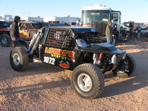 1990 Ford Bronco - black tube-chassis off-road race car numbered 1072 with Livewire Energy graphics sits on packed dirt amid spectators and trailers.