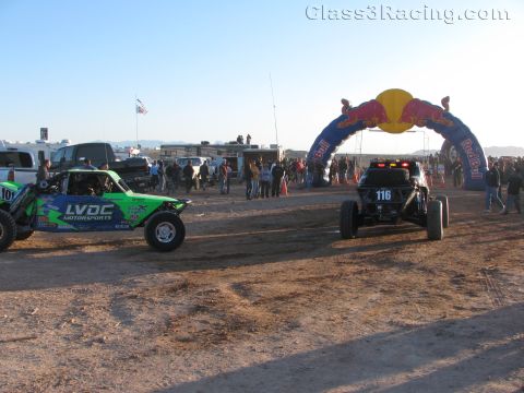 1990 Ford Bronco - green LVDC Motorsports buggy and car number 116 stage near a Red Bull inflatable arch in a dusty desert race paddock.