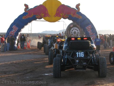 1990 Ford Bronco - line of off-road race cars including number 116 stages beneath a Red Bull inflatable arch as dust rises at a desert start line.