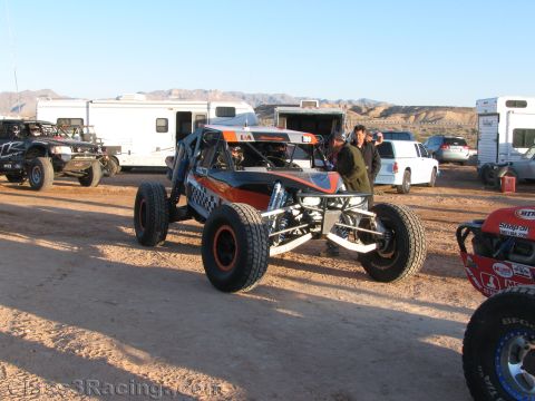 1990 Ford Bronco - orange and gray tube-chassis off-road buggy sits in a dirt pit area surrounded by RVs and trailers under desert mountains.