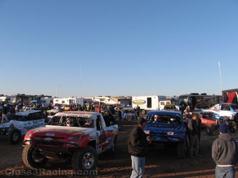 1990 Ford Bronco - Several off-road race trucks and buggies, including a red and a blue truck, sit in a dirt staging area with RVs and people at dawn.