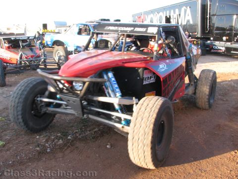 1990 Ford Bronco - red tube-chassis off-road race buggy with knobby tires sits on red dirt beside a Yokohama-branded transporter and other cars.