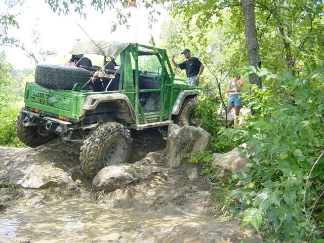 Green off-road vehicle with a spare tire on the back, navigating a rocky, muddy trail with two people standing nearby