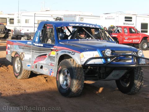 1990 Ford Bronco - blue and gray off-road prerunner race truck with Western livery and a driver inside sits on dirt among RVs.