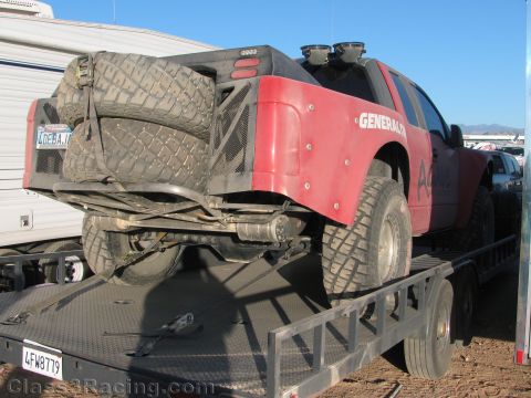1990 Ford Bronco - red and black General Tire off-road race truck sits strapped to a flatbed trailer with a muddy spare mounted at the rear.
