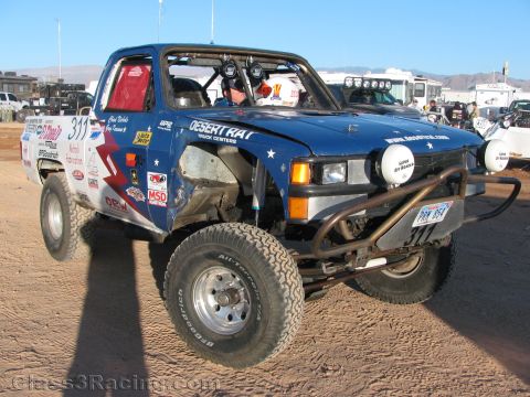 1990 Ford Bronco - blue off-road race truck number 311 with Desert Rat and BFGoodrich graphics and front light pods sits on dirt with a driver aboard.