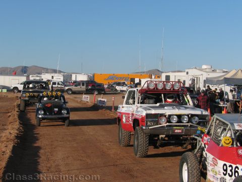 1990 Ford Bronco - black buggy and a white off-road truck move along a dusty paddock road lined with RVs, tents and other race cars.
