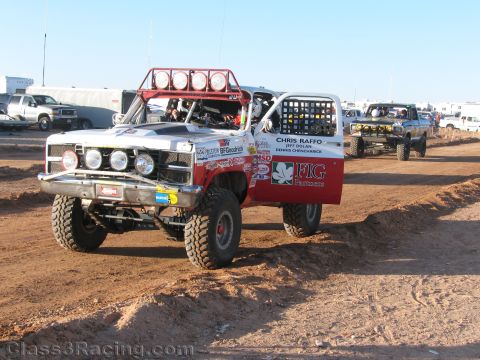 1990 Ford Bronco - white and red off-road race truck with a four-light roof bar and Chris Raffo team graphics sits on dirt among trailers.
