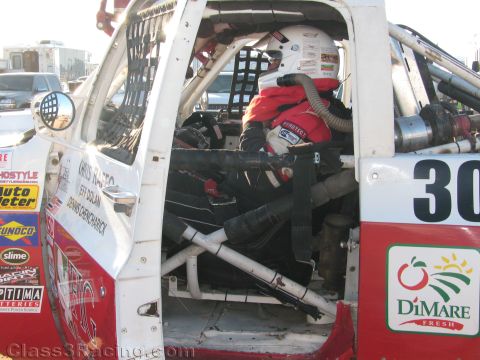 1990 Ford Bronco - driver in a red fire suit and white helmet sits strapped inside the roll cage of a white and red off-road race truck.