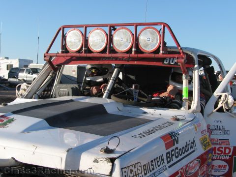 1990 Ford Bronco - View over the hood and roof light bar of a white off-road race truck with crew seated in the caged cockpit at a desert event.