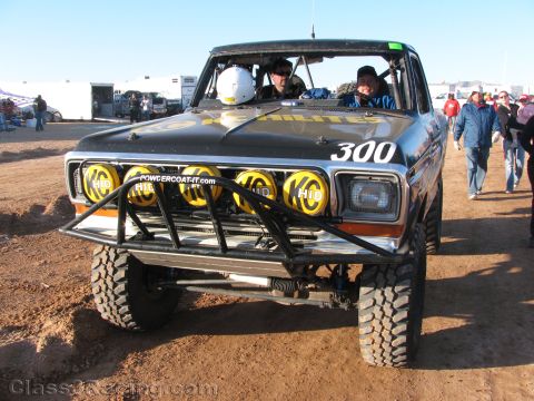 1990 Ford Bronco - black classic Ford off-road race truck numbered 300 with yellow Hella light pods and a tube bumper sits on dirt with crew aboard.