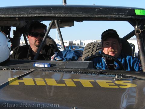 1990 Ford Bronco - Two men, one in sunglasses and one in a cap, sit in the cockpit of a black off-road race truck marked HiLites on the hood.