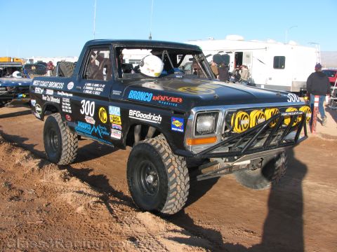 1990 Ford Bronco - black classic Ford off-road race truck numbered 300 with West Coast Broncos and KC light graphics sits on dirt among RVs.
