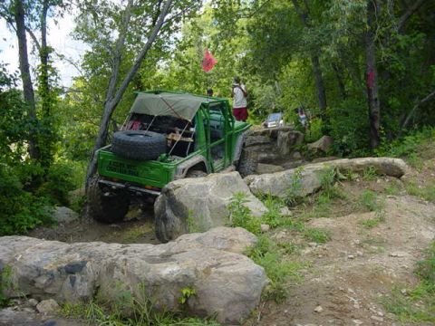 1992 Ford Bronco - A modified green off-road vehicle tackles a challenging rocky terrain with trees and foliage surrounding it.