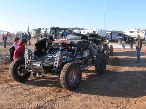 1990 Ford Bronco - black two-seat tube-chassis off-road race buggy numbered 1597 stages on dirt as crew and spectators stand nearby under blue sky.